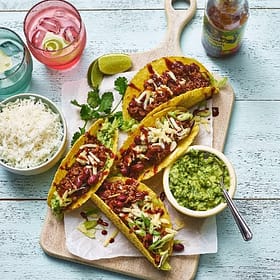 Fresh tacos with seasoned meat, cheese, and vegetables served on a wooden platter with guacamole, rice, and beverages, showcasing Cedar Works' fresh and flavorful food offerings.