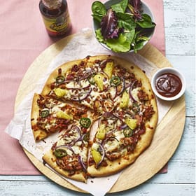 Fresh wood-fired vegetable pizza with jalapeños, pickles, and red onions on a rustic wooden serving board with a side salad and barbecue sauce.