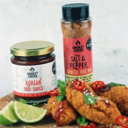 Smoky Carter Korean BBQ sauce and Chinese BBQ rub jars with crispy fried chicken and lime wedges on a rustic surface, showcasing premium BBQ seasoning products by Cedar Works.