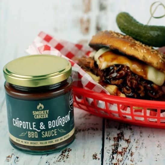 Savory BBQ sandwich with chipotle bourbon sauce in a red basket, featuring a dill pickle on top, served alongside a jar of Smokey Caker BBQ sauce. Perfect for grilled meats and outdoor cookouts.