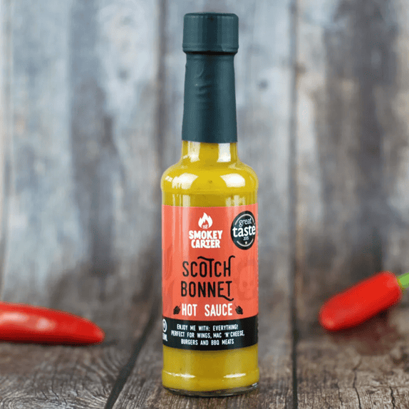 A bottle of Smokey Carter Scotch Bonnet hot sauce on a rustic wooden surface, surrounded by red chili peppers, emphasizing bold flavor and premium quality.