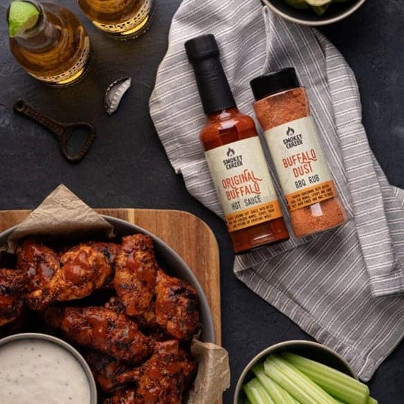 Smokey Chicken Original Buffalo Hot Sauce and Buffalo Dust BBQ Rub with grilled chicken wings, celery sticks, and dipping sauce on a dark table for flavorful BBQ meals.