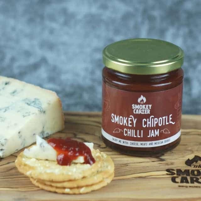 Smoky chipotle chili jam jar with cheese and crackers on wooden board, artisanal condiment, gourmet pairing, spicy sweet flavor, Cedar Works NZ.
