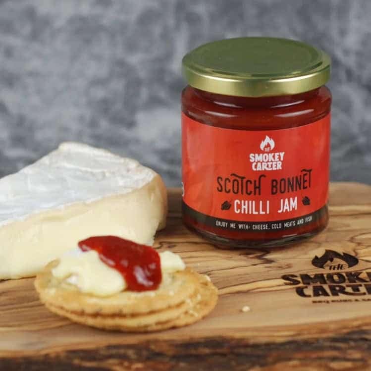 Scotch Bonnet chili jam jar with cheese and cracker on wooden board, vibrant condiment from Smokey Carter, ideal for cheese pairings and spicy snacks, featuring quality ingredients and bold flavors.